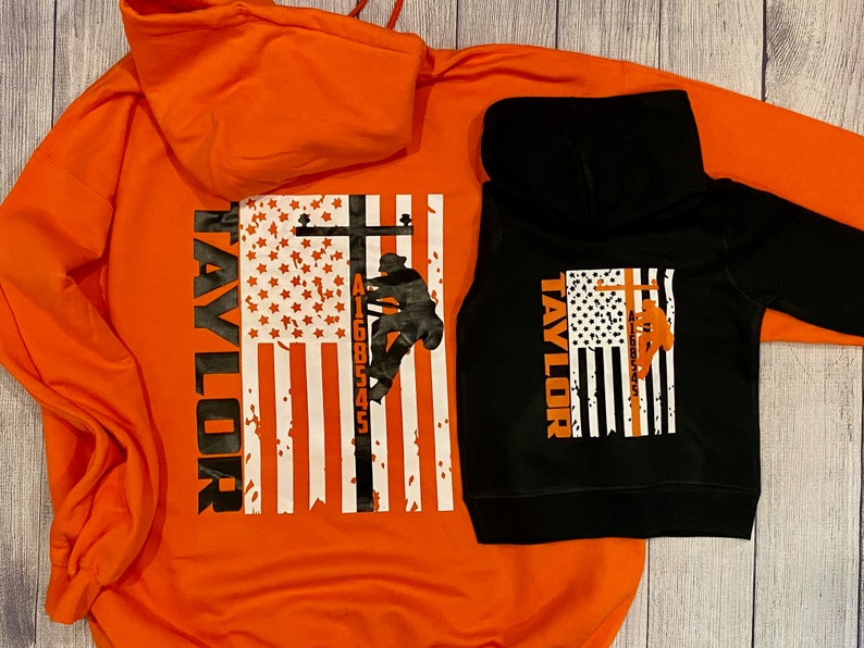 May include: Two hooded sweatshirts, one orange and one black, with a graphic of a lineman climbing a utility pole in front of an American flag. The text "TAYLOR" is printed vertically on the left side of the flag. The number "A1600545" is printed on the pole.