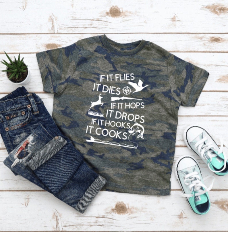 May include: A camouflage-patterned t-shirt with the text "IF IT FLIES IT DIES, IF IT HOPS IT DROPS, IF IT HOOKS IT COOKS" in white. The shirt features hunting and fishing icons.
