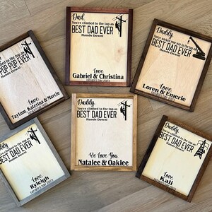 May include: A collection of six wooden signs with a brown wood frame. Each sign features a silhouette of a person working on a power line and the text "You've climbed to the top as BEST DAD EVER Hands Down!" with a different name and date for each sign.