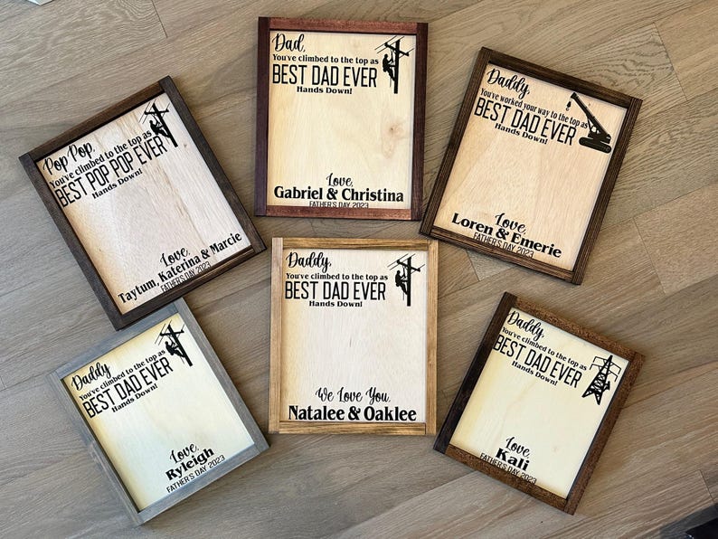 May include: A collection of six wooden signs with a brown wood frame. Each sign features a silhouette of a person working on a power line and the text "You've climbed to the top as BEST DAD EVER Hands Down!" with a different name and date for each sign.