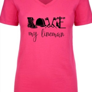 May include: Pink v-neck t-shirt with a black graphic that says "LOVE my lineman" and includes a boot, hard hat, pliers, and a utility pole.