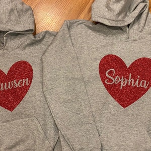 K&ouml;nnte beinhalten: Zwei graue Kapuzenpullover mit roten Glitzerherzen. Ein Herz sagt "Dawson" und das andere sagt "Sophia".