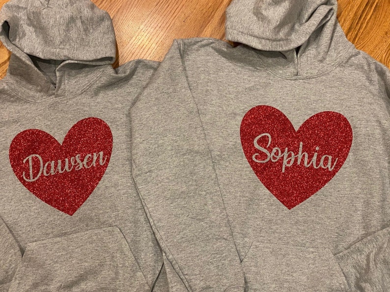 K&ouml;nnte beinhalten: Zwei graue Kapuzenpullover mit roten Glitzerherzen. Ein Herz sagt "Dawson" und das andere sagt "Sophia".