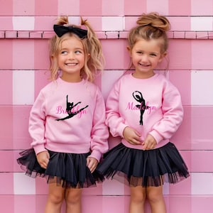Puede incluir: Dos niñas pequeñas con sudaderas rosas con diseños de bailarinas negras y nombres, combinadas con faldas de tul negras. Una sudadera dice "Brooklyn" y la otra "Madeleyn". Sonríen frente a un fondo rosa a rayas.