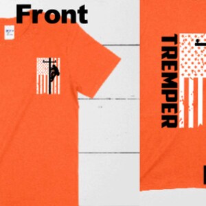 May include: Orange t-shirt with a small American flag design featuring a lineman on a utility pole on the front. The back of the shirt has a larger American flag design with the word "TREMPED" printed vertically on the left side.