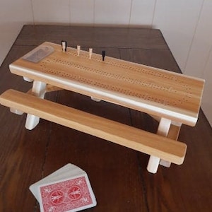 May include: A wooden cribbage board game set with a wooden picnic table design. The board has a white painted surface with holes for pegs. The set includes a deck of playing cards and pegs.