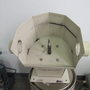 May include: A beige metal machine with an octagonal top, cylindrical tubes, and a central metal rod. The machine sits atop a metal drum with a brown paper exterior. The machine is likely used for industrial purposes.