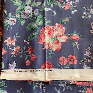 May include: Blue fabric with a floral print featuring pink, red, and white flowers. The fabric has a white label with the text "Cranston Print Works Company".