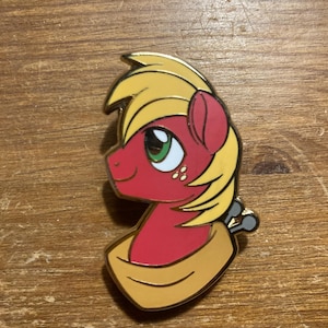 May include: A red and yellow enamel pin featuring a cartoon pony with a yellow mane and tail. The pony is wearing a gold scarf and has a green eye. The pin has a gold finish and a silver back.