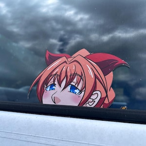 Puede incluir: Una pegatina con un personaje de estilo anime con pelo naranja y ojos azules. Se ve la cabeza del personaje, con un rubor en las mejillas y un lazo rojo. La pegatina está colocada en la ventana de un coche, con un cielo nublado de fondo.