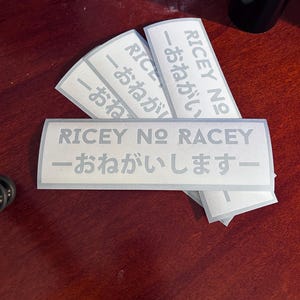 May include: Four white stickers with the text "RICEY NO RACEY - おねがいしますー" in Japanese and English. The stickers are stacked on top of each other.