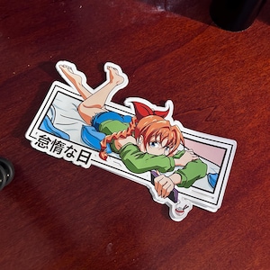 May include: A sticker featuring an anime-style illustration of a woman with red hair, wearing a green sweater and blue jeans, lying on a bed with a book. The sticker has a white border and the text "感情な日" in Japanese.