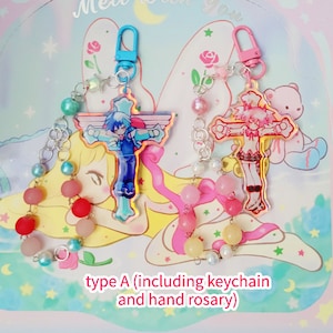 May include: Two colorful anime character cross charms with silver chains and beaded hand rosaries. The charms are attached to blue and pink keychains. The text "type A (including keychain and hand rosary)" is at the bottom of the image.