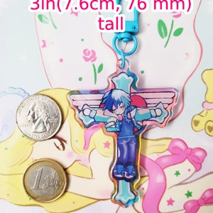 May include: A blue and pink iridescent charm featuring a character with blue hair and a blue and white outfit. The charm is shaped like a cross and has wings on the sides. The charm is attached to a blue keychain with a silver clasp. The charm is 3 inches (7.6 cm) tall.