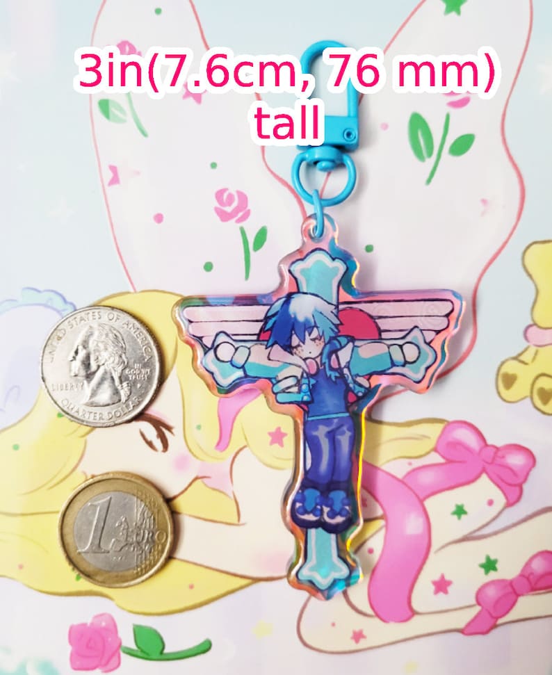 May include: A blue and pink iridescent charm featuring a character with blue hair and a blue and white outfit. The charm is shaped like a cross and has wings on the sides. The charm is attached to a blue keychain with a silver clasp. The charm is 3 inches (7.6 cm) tall.