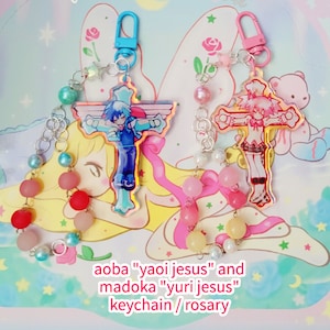 May include: Two colorful anime character keychains with a cross design. The keychains are attached to a silver chain with pink, blue, and white beads. The text "aoba "yaoi jesus" and madoka "yuri jesus" keychain / rosary" is visible at the bottom of the image.