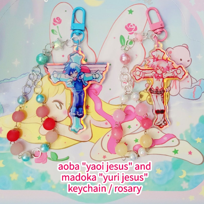 May include: Two colorful anime character keychains with a cross design. The keychains are attached to a silver chain with pink, blue, and white beads. The text "aoba "yaoi jesus" and madoka "yuri jesus" keychain / rosary" is visible at the bottom of the image.
