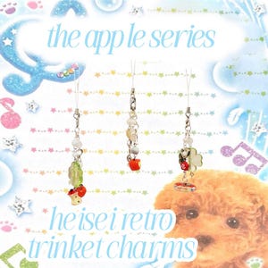 May include: Three decorative trinket charms with silver chains and various beads. Each charm features a different design, including a red apple, a green cactus with red apples, and a four-leaf clover with a ladybug. The text "the apple series" and "heisei retro trinket charms" are visible.