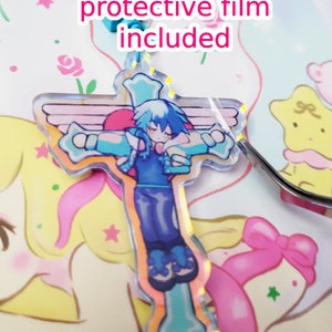 May include: A blue and white anime character with wings is depicted on a clear acrylic charm in the shape of a cross. The charm has a protective film on it. The charm is on a white surface with a pink and yellow background.