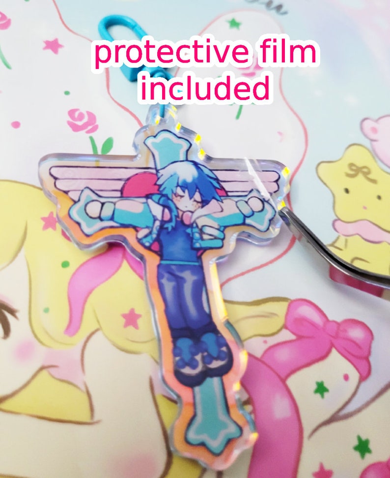 May include: A blue and white anime character with wings is depicted on a clear acrylic charm in the shape of a cross. The charm has a protective film on it. The charm is on a white surface with a pink and yellow background.