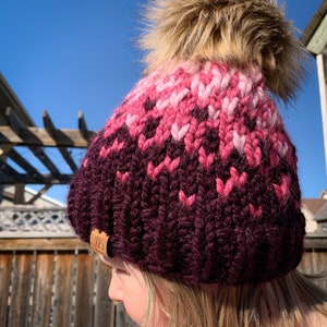Puede incluir: Un gorro de punto rosa y burdeos con un pomp&oacute;n de piel sint&eacute;tica. El gorro tiene una etiqueta de cuero marr&oacute;n con la palabra "LOOPS" en ella.