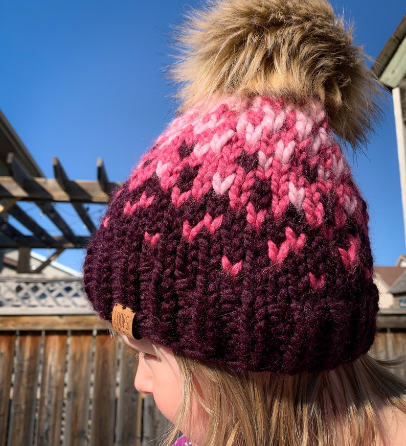 Puede incluir: Un gorro de punto rosa y burdeos con un pomp&oacute;n de piel sint&eacute;tica. El gorro tiene una etiqueta de cuero marr&oacute;n con la palabra "LOOPS" en ella.