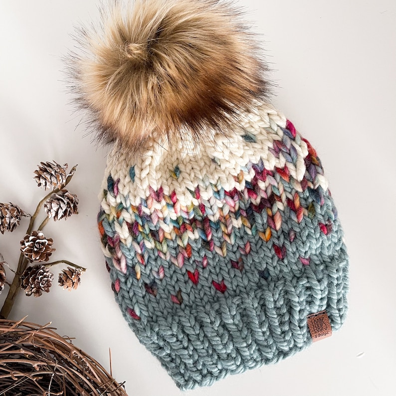 Puede incluir: Un gorro de punto azul, blanco y multicolor con un pomp&oacute;n marr&oacute;n esponjoso. El gorro tiene una etiqueta de cuero marr&oacute;n con la palabra "LOOP" en ella.