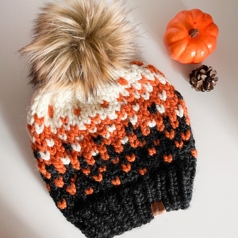 Puede incluir: Un gorro de punto negro, naranja y blanco con un pomp&oacute;n marr&oacute;n esponjoso en la parte superior.