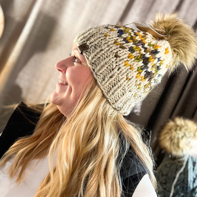 Puede incluir: Un gorro de punto de color crema con un pomp&oacute;n en la parte superior. El gorro tiene un patr&oacute;n geom&eacute;trico colorido en tonos de amarillo, azul y verde.