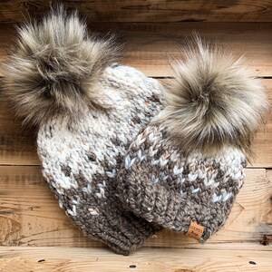 Puede incluir: Dos gorros de punto con pompones de piel sint&eacute;tica. Los gorros son marrones y blancos con un patr&oacute;n moteado. Los gorros tienen una etiqueta de cuero marr&oacute;n con el texto "Loops & Things" en ellos.