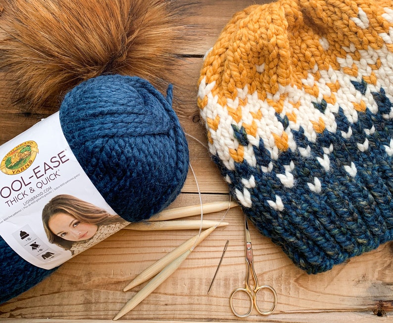 Puede incluir: Un gorro de punto azul y blanco con un patr&oacute;n amarillo y blanco. El gorro est&aacute; hecho con hilo Lion Brand Cool Ease Thick & Quick. El hilo tambi&eacute;n se muestra en la imagen, junto con agujas de tejer y tijeras.