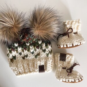 Puede incluir: Un gorro de punto blanco con dos pompones de piel sint&eacute;tica marr&oacute;n y una etiqueta marr&oacute;n "LOOPS & more". El gorro se combina con un par de botines de punto blanco con ribetes marrones y una etiqueta marr&oacute;n "LOOPS & more".