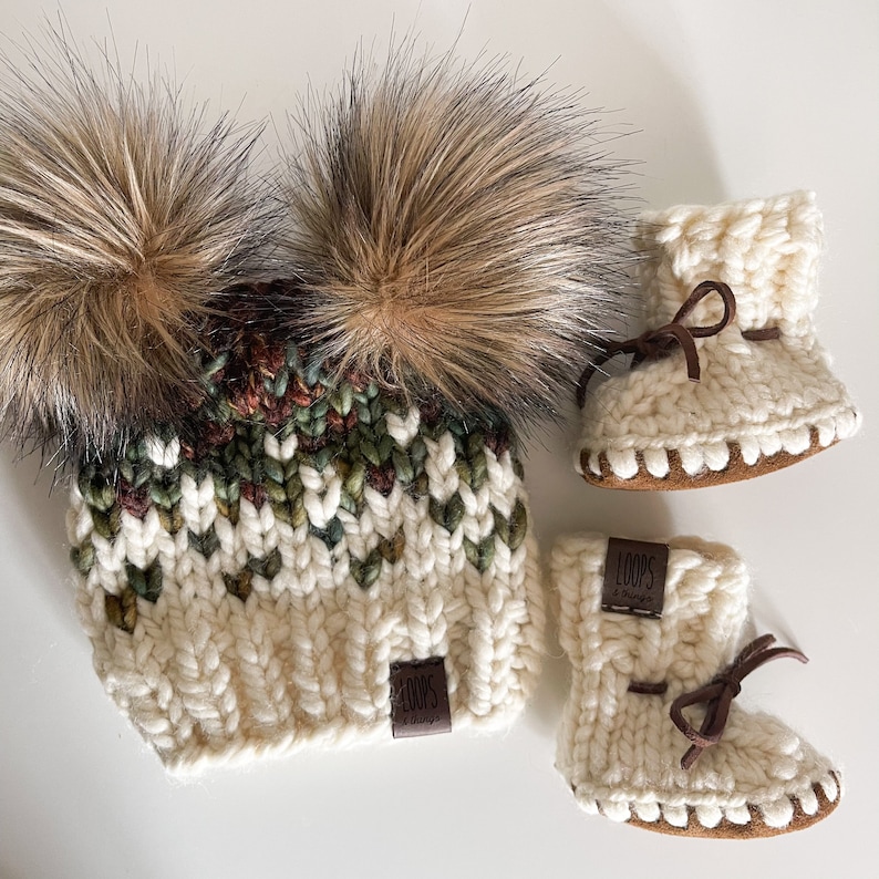 Puede incluir: Un gorro de punto blanco con dos pompones de piel sint&eacute;tica marr&oacute;n y una etiqueta marr&oacute;n "LOOPS & more". El gorro se combina con un par de botines de punto blanco con ribetes marrones y una etiqueta marr&oacute;n "LOOPS & more".