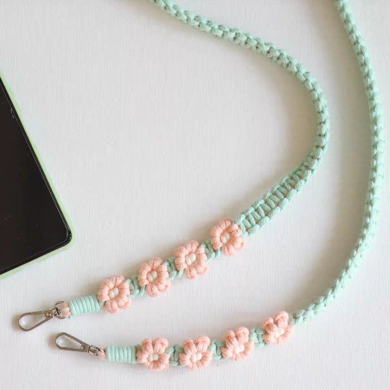 May include: A light green and pink phone strap with crocheted flower accents. The strap has two metal clasps.