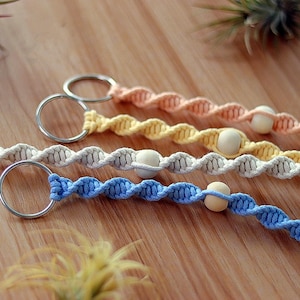 May include: Four colorful macrame keychains with wooden beads. The keychains are in shades of blue, beige, yellow, and peach. Each keychain has a silver key ring.