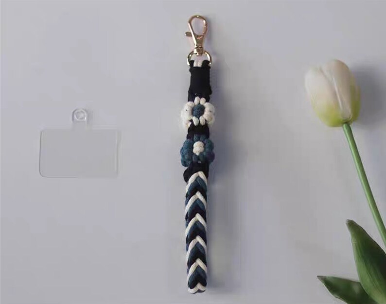 Pu&ograve; includere: Un laccio intrecciato bianco e nero con un portachiavi dorato e tre ciondoli a forma di fiore blu e bianco. Il laccio &egrave; appeso su uno sfondo bianco.