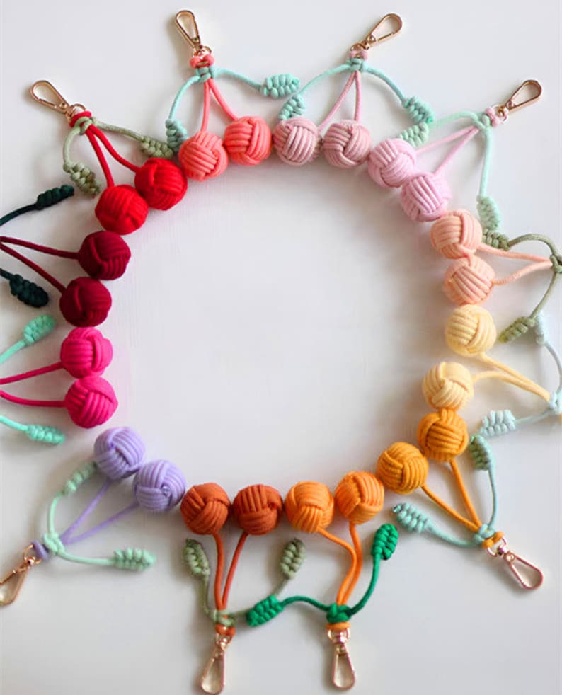 Op de afbeelding: Een regenboogassortiment van kleurrijke geknoopte sleutelhangers met gouden metalen sluitingen. Elke sleutelhanger heeft een paar geknoopte ballen en een groene blaadjescharme.