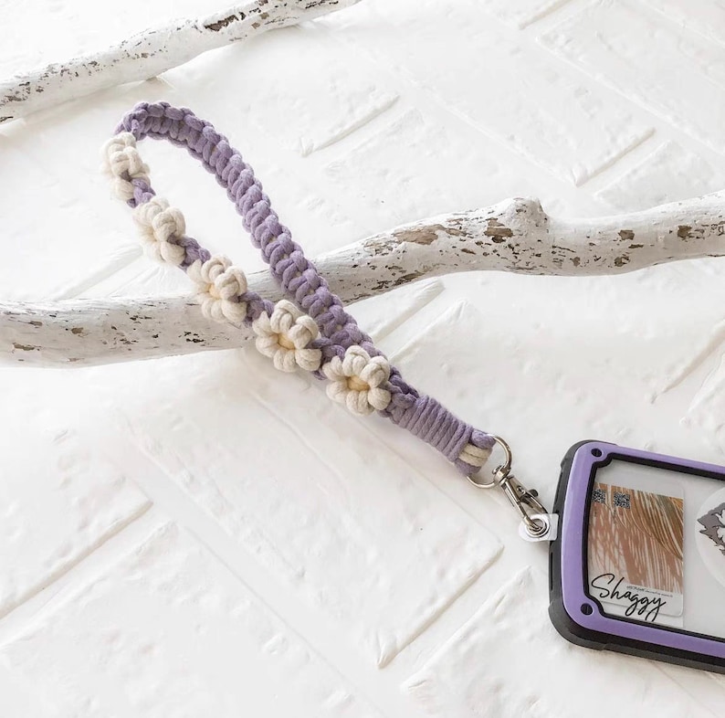 May include: A purple and white crocheted phone strap with a daisy flower design. The strap has a silver metal clip attached to it.