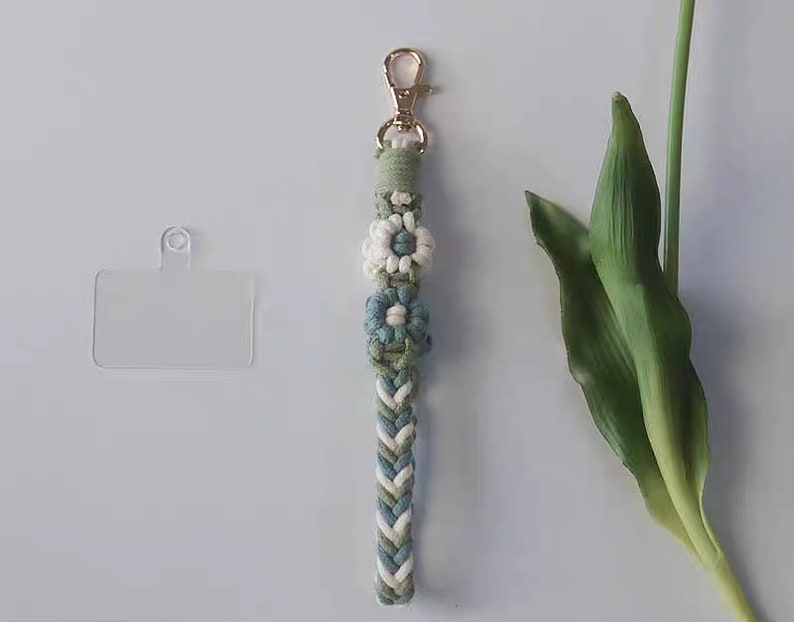 Pu&ograve; includere: Un portachiavi intrecciato verde, bianco e blu con una chiusura dorata e tre ciondoli a forma di fiore bianco e blu. Il portachiavi &egrave; appeso a un porta carte in plastica trasparente.