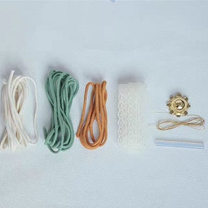 K&ouml;nnte beinhalten: Ein Set an Bastelmaterialien f&uuml;r die Herstellung einer Makramee-Wandbeh&auml;nge. Das Set enth&auml;lt wei&szlig;e, gr&uuml;ne und orangefarbene Schn&uuml;re, wei&szlig;e Spitzenborte, einen goldenen Metallring, goldenen Faden und einen durchsichtigen Klebestift.