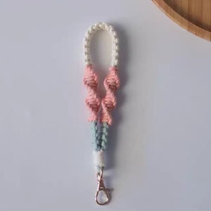 Pode incluir: Um chaveiro de macram&eacute; rosa, branco e azul com um aro de chave prateado.