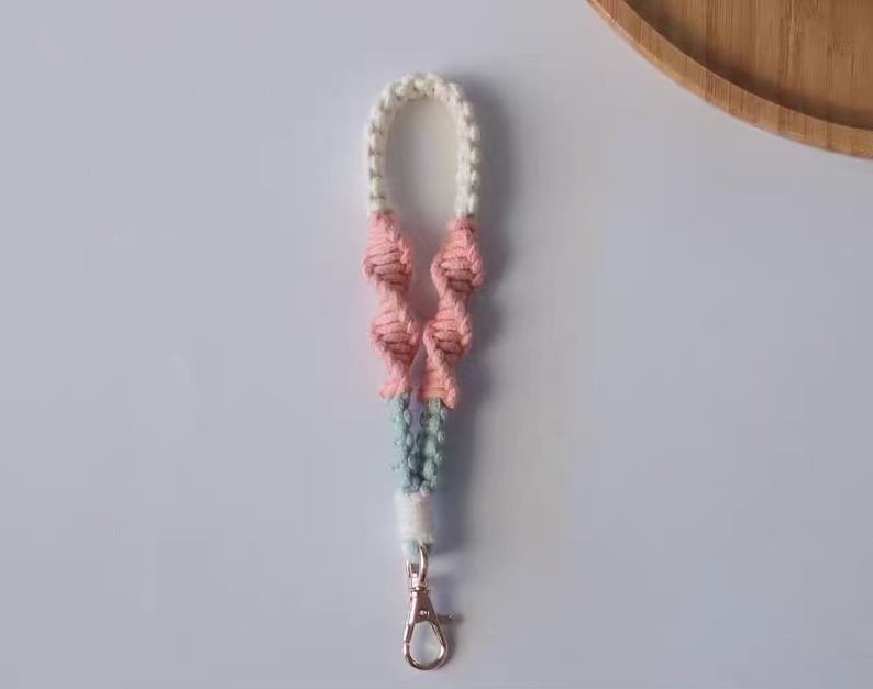 Pode incluir: Um chaveiro de macram&eacute; rosa, branco e azul com um aro de chave prateado.
