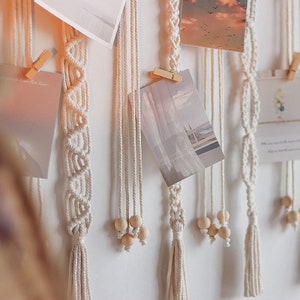 Op de afbeelding: Een witte macram&eacute; wandhanger met houten kralen en kwastjes. Foto's worden met wasknijpers aan de wandhanger bevestigd.