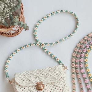 May include: A cream-colored macrame bag with a gold clasp and a daisy chain strap. The strap features white, yellow, and green details. Additional daisy chain straps in pink, yellow, and peach are visible.