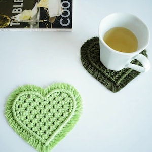 以下が含まれることがあります： フリンジの縁取りが施された、緑とオリーブグリーンの2つのハート型のクロシェコースター。オリーブグリーンのコースターの上に、お茶が入った白いマグカップが置かれています。