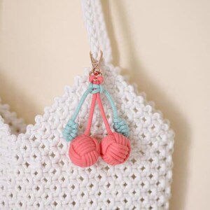 Op de afbeelding: Een witte gehaakte tas met een roze en blauwe kers sleutelhanger. De sleutelhanger heeft twee roze pompoenen en blauwe touwtjes.