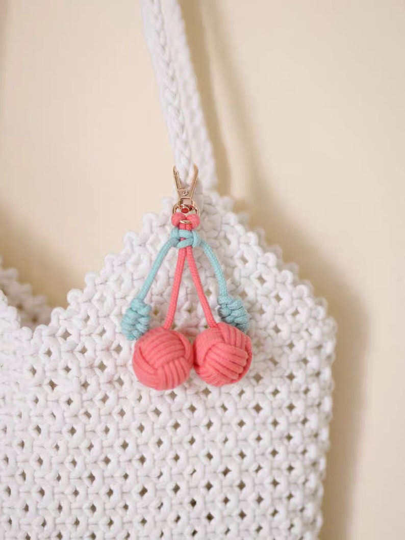 Op de afbeelding: Een witte gehaakte tas met een roze en blauwe kers sleutelhanger. De sleutelhanger heeft twee roze pompoenen en blauwe touwtjes.
