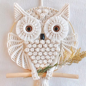 May include: White macrame owl wall hanging with a wooden dowel and a sprig of dried flowers. The owl has large, round eyes with brown pupils.