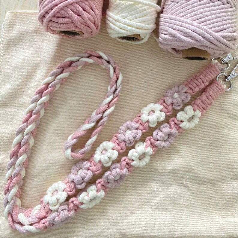 May include: A pink and white macrame cord necklace with flower accents. The necklace is made with two strands of cord that are braided together and then knotted into a flower pattern.