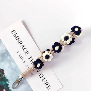 Może przedstawiać: Brelok do kluczy z makramą w kolorze beżowym i granatowym z kwiatowym wzorem. Brelok ma srebrny metalowy zamek.